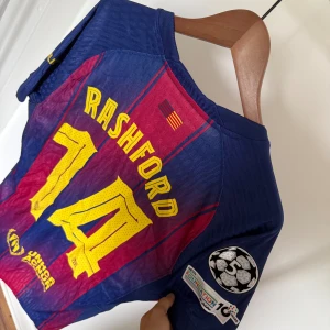 Barcelona 25/26 Rashford #14 Nike tröja M - Säljer en officiell FC Barcelona matchtröja med Rashford och nummer 14 på ryggen. Tröjan är randig i mörkblått och vinrött med gula detaljer, Nike-logga och Spotify sponsor på bröstet. Materialet är lätt och andas, perfekt för fotboll. Champions League-märke på ärmen.