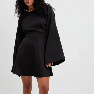 Svart kort klänning med öppen rygg - Supersnygg svart kort klänning med vida ärmar och öppen rygg som knyts med tunna band. Klänningen har en lätt glansig finish och markerad midja, vilket ger en elegant och trendig look. Perfekt för dig som vill sticka ut med stilrena detaljer. Helt ny med prislapp kvar, aldrig använd !