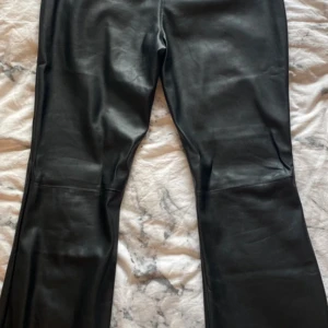 Svarta bootcut byxor i skinnimitation Nelly - Snygga svarta byxor från Nelly i skinnimitation med bootcut-modell. Byxorna har låg midja, två knappar framtill och raka ben som slutar i en lätt utsvängning. Perfekta för dig som vill ha en edgy och trendig look. Nya använda 1 gång.