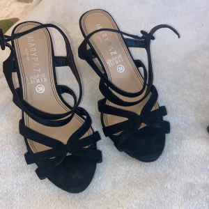 Svarta sandaler med klack från Marypaz - Svarta sandaler från Marypaz med snygga remmar och öppen tå. Skorna har hög klack och spänne runt vristen. Tillverkade i mockaliknande material som ger en elegant känsla. Perfekta för dig som vill ha en stilren och trendig look.