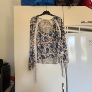 Odd Molly blommig blus med spets - Supersöt blommig blus från Odd Molly i beige med blått och vinrött mönster. Blusen har v-ringning, långa ärmar, virkade spetsdetaljer och knytband framtill. Perfekt för dig som gillar boho-vibe och unika detaljer. Storlek 1 som enligt deras storleksguide motsvarar en storlek S. Men den kan vara något rymlig i storlek. Mycket fint skick, jag hittar inga synliga anmärkningar eller defekter. 