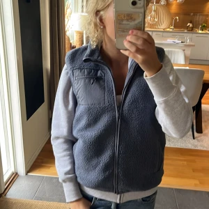 Mörkblå/grå fleeceväst med dragkedja - Säljer en mörkgrå fleeceväst med hög krage och hel dragkedja framtill. Västen har en snygg quiltad ficka med dragkedja på bröstet och är perfekt att slänga över en hoodie. Passformen är normal och materialet är mjuk fleece. Aldrig använd!