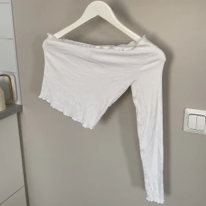 Vit offshoulder topp från ASOS - Säljer en vit ribbad offshoulder topp från ASOS i storlek 36. Toppen har en croppad passform, lång ärm på ena sidan och vågig kant nertill och upptill. Perfekt för dig som gillar trendiga och unika plagg med snygg passform.