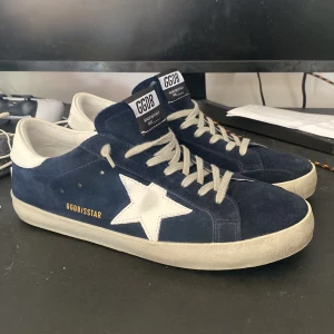 Golden Goose Superstar sneakers blå mocka - Golden Goose Superstar sneakers i mörkblå mocka med vit stjärndetalj på sidan och vit hälkappa. Skorna har grå snörning, platt sula och klassisk rund tå. Märkeslogga bak och på plösen. Perfekt för dig som gillar streetstyle och premiumkänsla. (Priset kan diskuteras vid snabb affär) Har använt dessa 2 gånger, tyckte inte det matcha min stil så beslutade mig för att sälja de. 