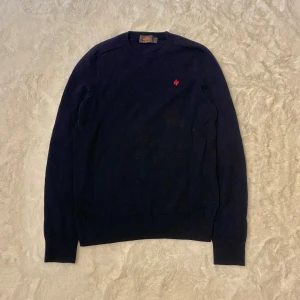 Mörkblå merinoullströja från Morris S - Mörkblå stickad tröja från Morris i 100% merinoull. Klassisk modell med rund halsringning och lång ärm. Broderad röd lilja på bröstet ger en snygg detalj. Perfekt för dig som gillar stilrena och tidlösa plagg.