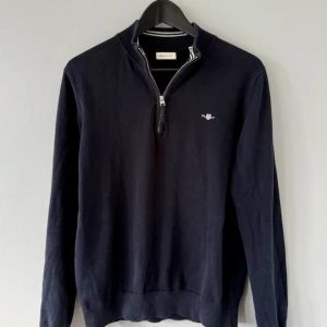 Mörkblå half zip tröja från Gant - Snygg mörkblå tröja från Gant med halv dragkedja och broderad logga på bröstet. Tröjan har ribbade muddar och hög krage, perfekt för lager-på-lager. Tillverkad i mjuk bomull som känns skön mot huden.