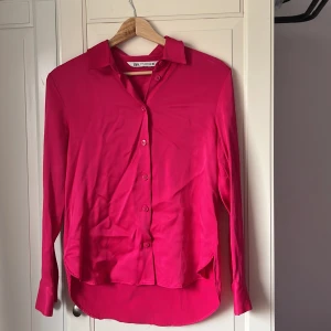Ceriserosa skjorta från Zara XS - Säljer en ceriserosa skjorta från Zara i storlek XS. Skjortan har klassisk krage, knäppning framtill och långa ärmar med manschett och knapp. Materialet är mjukt och har en lätt glansig finish som ger en snygg look.