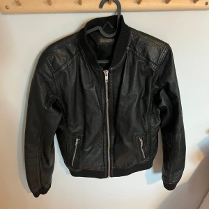 Svart skinn bomberjacka Rockandblue - Snygg svart bomberjacka från Rockandblue i skinn med ribbad krage och muddar. Jackan har två fickor med dragkedja och en silverfärgad zip framtill. Perfekt till hösten. Snygg passform. Storlek 40 men passar 36/38 om man vill ha en lite oversized look.