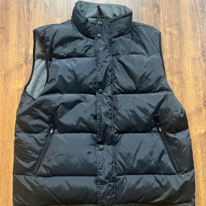 Svart dunväst från Grizzly XXL - Svart dunväst från Grizzly i storlek XXL. Västen har quiltad design, hög krage och två dragkedjeförsedda fickor framtill. Insidan är fodrad med mjuk grå fleece för extra värme. Perfekt för lager-på-lager-stil under kyliga dagar.