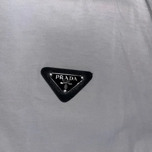  Prada tshirt - Vit t-shirt från Prada med klassisk rund hals och korta ärmar. På bröstet sitter en svart, triangelformad Prada-logga i metall och läder. T-shirten är i mjuk bomull och har en clean, stilren look som passar perfekt till streetwear eller casual outfits.