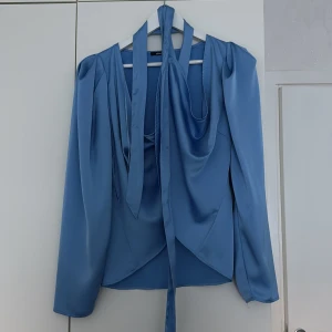 Blå omlottblus i satin från Gina Tricot - Supersnygg blå omlottblus i  satinmaterial från Gina Tricot. Blusen har långa ärmar, markerade axlar och knytband i midjan som ger en riktigt chic siluett. Djup v-ringning framtill och lätt puff vid axlarna för extra stilpoäng.