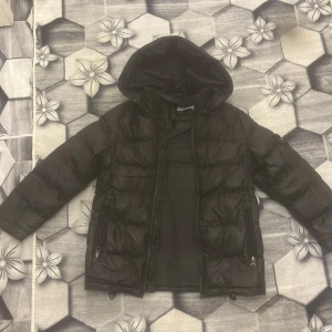 Svart pufferjacka från Stone Island - Säljer en svart pufferjacka från Stone Island med huva och klassisk patch på vänster ärm. Jackan har dragkedja framtill, två fickor med dragkedja och quiltad design. Perfekt för kalla dagar och riktigt snygg streetstil.