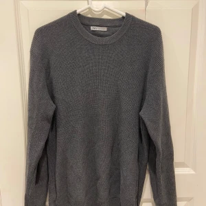 Grå stickad tröja från Zara - En stilren grå stickad tröja från Zara i storlek S. Tröjan har rund halsringning, ribbade muddar och är långärmad. Perfekt för lager-på-lager och enkel att matcha med jeans eller kjol. Passar också M