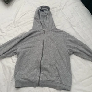 Grå hoodie med dragkedja från H&M - En enkel och chill grå hoodie från H&M med hel dragkedja framtill, huva och två stora fickor. Perfekt för att slänga på sig när det är lite kyligt. Mjuk bomullskänsla och långärmad för extra komfort.