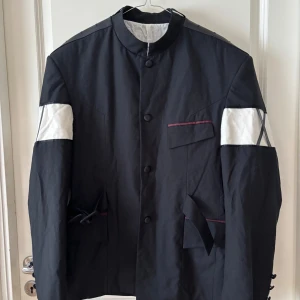 Svart kavaj med vita detaljer och bälte - Unik svart kavaj med ståkrage, vita paneler på ärmarna och snygga bältesdetaljer i midjan. Jackan har knappar framtill, fickor med vinröda detaljer och tre knappar vid ärmslutet. Materialet känns som bomull eller polyester och designen är riktigt edgy. Spänne på ryggen.