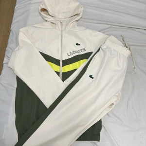 Vit hoodie från Lacoste med huva - Säljer en snygg vit hoodie från Lacoste med dragkedja och huva. Tröjan har gröna och gula detaljer på bröstet samt broderad Lacoste-logga. Perfekt för dig som gillar sportig och clean stil. Materialet känns mjukt och lätt, och passformen är normal.