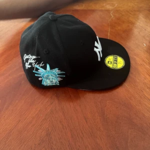 Svart New Era Yankees 59FIFTY keps - Snygg svart 59FIFTY keps från New Era med New York Yankees-logga framtill. Broderad frihetsgudinna och texten 'Greetings from New York' på sidan. Klassisk rak skärm och broderade detaljer i vitt och blått. Perfekt för dig som vill ha streetstyle med NYC-vibe.                                                         Beställde en storlek för stort😔