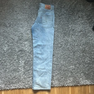 Levi's 501 ljusblå jeans - Klassiska Levi's 501 jeans i ljusblå tvätt med raka ben och hög midja. Jeansen har den ikoniska läderpatchen bak i midjan och röd Levi's-tagg på bakfickan. Perfekta för en avslappnad och tidlös stil.