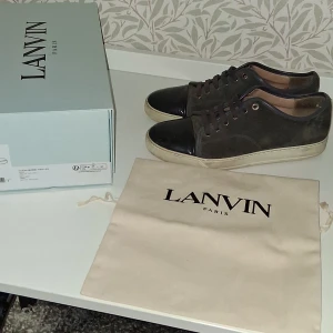 Lanvin sneakers  - Snygga sneakers från Lanvin i mörkgrå mocka med svarta skinn-detaljer på tån och mörka snören. Skorna har vit sula och diskreta metalldetaljer vid snörningen. Kommer med originalkartong och dustbag. Perfekta för dig som gillar stilrena och exklusiva sneakers. Finns några få skador som på hälen på ena skon och även små andra defekter som man kan få bild på om man frågar! Köpte skorna för cirka 5000kr så tycker ändå att 2600kr är ett rimligt pris, hör av er vid fler frågor!