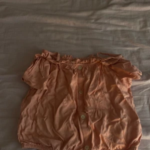 Persikofärgad blus från LC Waikiki - Söt persikofärgad blus från LC Waikiki med korta ärmar och offshoulder-modell. Blusen har knappar framtill, resår vid halsringning och nederkant samt volangdetalj upptill. Perfekt för en somrig och avslappnad look.