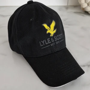 Svart keps från Lyle & Scott - Svart keps med böjd skärm från Lyle & Scott. Framtill finns en gul broderad fågel och logotypen i grått. Skärmen har en vit kant som ger en snygg kontrast. Kepsen är tillverkad i bomull och har klassisk passform med justerbar rem baktill.