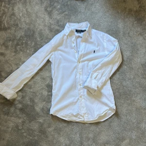 Vit skjorta från Polo Ralph Lauren XS - Klassisk vit skjorta från Polo Ralph Lauren i bomull, custom fit. Skjortan har lång ärm, knappar framtill och den ikoniska broderade loggan på bröstet. Perfekt till jeans eller chinos för en clean och stilren look. Använd en gång på skolavslutning 