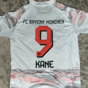 Bayern München Kane #9 Adidas tröja M - FC Bayern München fotbollströja med Kane och nummer 9 på ryggen. Adidas AEROREADY-modell i vitt med grå och röda grafiska detaljer, svarta ränder på axlarna och klubbmärke på bröstet. Slim fit och kortärmad, perfekt för fans.
