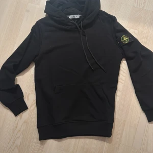 Svart hoodie från Stone Island - Svart hoodie från Stone Island med klassisk patch på ärmen. Tröjan har huva med snörning, magficka och långa ärmar. Perfekt för dig som gillar streetwear och vill ha en stilren look.