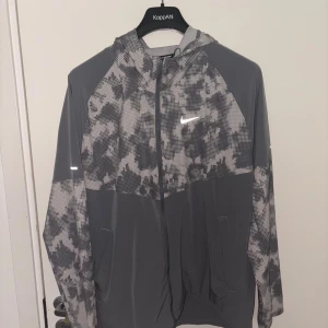 Grå mönstrad vindjacka från Nike - Snygg grå vindjacka från Nike med digitalt camo-mönster i ljusgrått och mörkgrått. Jackan har huva, hel dragkedja framtill och två fickor. Perfekt för träning eller när du vill ha en sportig look. Reflekterande Nike-logga på bröstet och detaljer på ärmarna.
