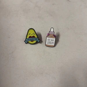Två färgglada pins med motiv - Säljer två coola pins: en med en söt avokado och texten 'Avo Good Time' i grönt och blått, samt en med en serumflaska och texten 'Self Love Serum' i beige, rosa och lila. Perfekta att fästa på jackan eller väskan för en personlig touch. 50 kr för båda och köparen står för frakten :)