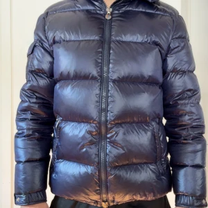 Mörkblå Moncler pufferjacka - Säljer en snygg mörkblå pufferjacka från Moncler med glansig finish och klassisk logga på fickan. Jackan har dragkedja framtill, långa ärmar och quiltad design som ger en cool och sportig vibe. Perfekt för kalla dagar när du vill hålla stilen. Pris går att diskutera😊😊