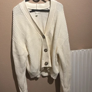 Vit stickad kofta med stora knappar - Säljer en vit stickad kofta från H&M med grov ribbad struktur och stora, snygga knappar framtill. Koftan har en djup v-ringning och långa ärmar med lite loose passform. Perfekt att slänga över en t-shirt för en chill look.
