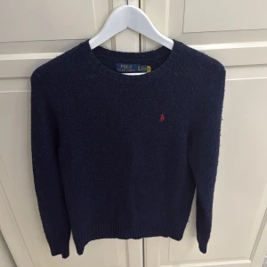 Mörkblå stickad tröja Polo Ralph Lauren - Mysig mörkblå stickad tröja från Polo Ralph Lauren i storlek XS. Klassisk modell med rund halsringning, ribbade muddar och diskret röd logga broderad på bröstet. Perfekt för lager-på-lager och chill stil.
