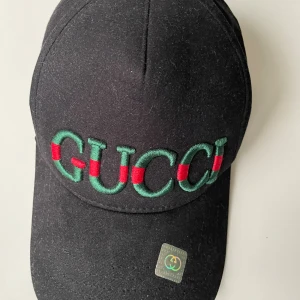 Svart Gucci keps med broderi - Svart keps från Gucci med böjd skärm och justerbar rem baktill. Framtill är märket broderat i grönt och rött, vilket ger en lyxig och ikonisk look. Kepsen är tillverkad i bomull och har en klassisk form som passar till streetwear och mode.