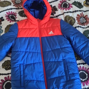 Blå och röd pufferjacka från Adidas - Snygg pufferjacka från Adidas i blått och rött med huva och dragkedja framtill. Jackan har quiltad design, två fickor och Adidas-logga på bröstet. Perfekt för kalla dagar när du vill hålla stilen och värmen.