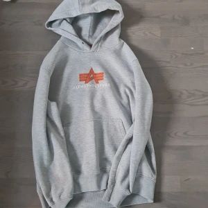 Grå hoodie Alpha Industries L - Säljer en grå hoodie från Alpha Industries i storlek L. Tröjan har en stor huva, känguruficka och ett snyggt orange logotryck på bröstet. Perfekt för chill dagar och streetstyle. Mjuk insida och klassisk passform.