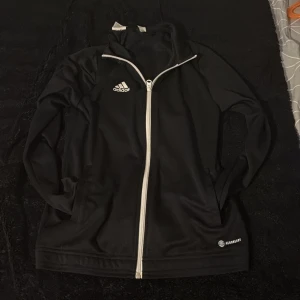 Svart Adidas ziptröja AEROREADY - Svart långärmad ziptröja från Adidas med vit dragkedja och logga på bröstet. Modellen är AEROREADY och har en sportig look, perfekt för träning eller chill. Tillverkad i polyester för en lätt och smidig känsla.