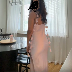 Ljusrosa långklänning med volangband - Superfin ljusrosa långklänning i satinliknande material med smala axelband och dekorativa volangband med små blommor längs axlarna. Klänningen har en snygg v-ringning och slits på sidan för extra stilpoäng. Oanvänd 