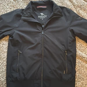 Svart jacka från Snoot Stockholm - Snygg svart jacka från Snoot Stockholm i storlek M. Jackan har dragkedja framtill, två fickor med dragkedja och hög krage. Materialet är slätt och lätt, perfekt för blåsiga dagar. Diskret Snoot-logga på ärmen och broderad logga på insidan. Säljer på grund av att den är för liten. Nypris  2500:- 