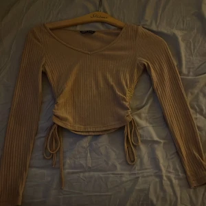 Ribbad croppad topp med snörning - Säljer en beige aktig långärmad topp från SHEIN med ribbad struktur och v-ringning. Toppen är croppad och har snygg snörning i sidorna som går att justera för en personlig passform. 