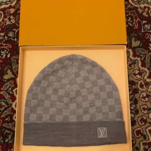 Grå Louis Vuitton rutig mössa - Exklusiv grå mössa från Louis Vuitton med klassiskt rutigt mönster och LV-logga framtill. Mössan är stickad och har en uppvikt kant för extra stilpoäng. Perfekt accessoar för dig som vill ha en lyxig och trendig look.