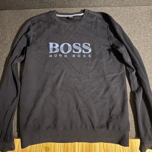 Mörk blå långärmad tröja Hugo Boss - Mörk blå  långärmad tröja från Hugo Boss med stort ljusblått BOSS-tryck på bröstet. Tröjan har rund hals och  perfekt för en clean och stilren look. Passar dig som gillar klassiska märken och enkel design.