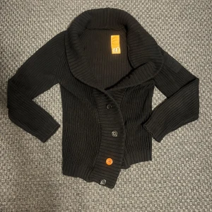 Svart stickad kofta från Mouli M - Svart ribbstickad kofta från Mouli med bred krage och fyra knappar framtill, varav en är orange för en cool detalj. Långärmad och i grovstickad bomull, perfekt för lager-på-lager. Modern Urban Lifestyle-märke på ärmen. Sitter rätt långt så passar runt m skulle jag säga! Inga defekter! 