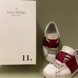 Valentino Garavani Open sneakers vit/röd - Snygga Valentino Garavani Open sneakers i vitt skinn med bred röd läderdetalj över mitten. Klassisk låg modell med rund tå, platt sula och snörning. Perfekta för dig som vill ha en lyxig och stilren sneaker med en unik touch.