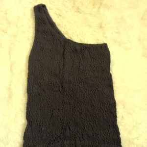 Svart oneshoulder topp med struktur - Säljer en svart topp med oneshoulder-design och snyggt strukturerat mönster i tyget. Toppen är ärmlös och har en cool asymmetrisk form som sticker ut. Perfekt för dig som vill ha något unikt och trendigt i garderoben.