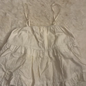 Vit volangig linnetopp med smala axelband - Supersöt vit linnetopp med smala axelband och flera lager volanger. Toppen är luftig och har en avslappnad passform, perfekt för varma dagar. Tillverkad i ett lätt material som känns skönt mot huden. Enkel och stilren design som är lätt att matcha.