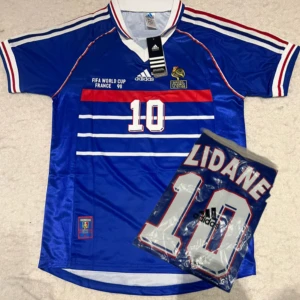Frankrike Zidane VM 98 tröja Adidas - Klassisk blå Frankrike fotbollströja från VM 1998 med Zidane och nummer 10 på ryggen. Adidas-logga, vita och röda detaljer samt horisontella ränder framtill. Tillverkad i glansigt syntetmaterial, kortärmad och med broderade märken.