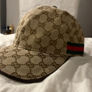 Gucci keps med GG-mönster och läderdetalj - Exklusiv keps från Gucci i beige och brunt GG-monogram, klassisk böjd skärm och justerbar rem i brunt läder. Sidan pryds av den ikoniska gröna och röda randen. Tillverkad i bomullscanvas med lyxiga detaljer och tydlig logotyp på insidan.