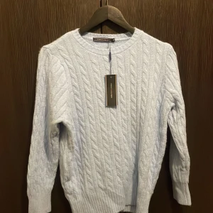 Ljusblå stickad tröja Tommy Hilfiger - Kabelstickad ljusblå tröja från Tommy Hilfiger med rund halsringning och långa ärmar. Tröjan har klassisk ribbad mudd vid ärmslut och nederkant, perfekt för lager-på-lager-stil. Snygg och enkel att matcha med jeans eller kjol.