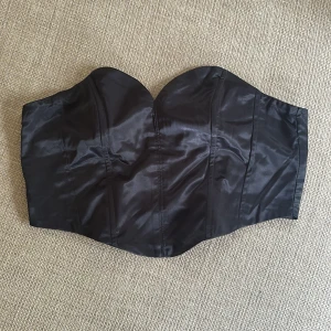 Svart satin korsett med dragkedja - Snygg svart korsett i glansigt satinmaterial med hjärtformad urringning och markerade sömmar. Toppen är axelbandslös och har en dragkedja bak för enkel på- och avtagning. Perfekt för att skapa en edgy och trendig look.